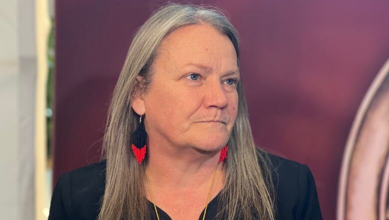 A woman with long, light-coloured hair and black/red beaded earrings is pictured looking at someone off-camera.