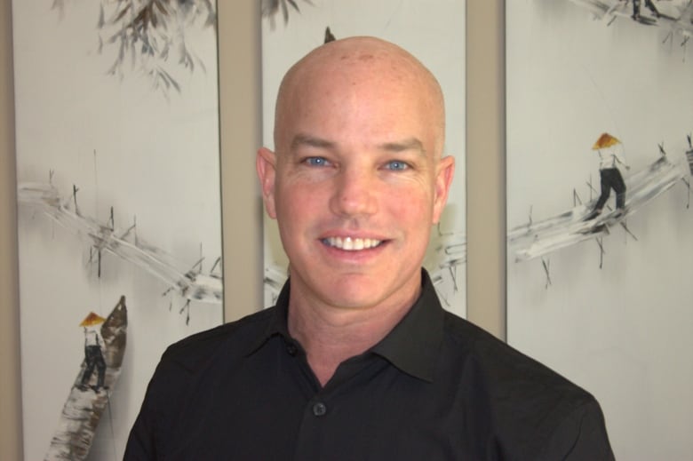 A bald man with blue eyes smiles for a photo. Behind him are pieces of art depitcing a man crossing a bridge and riding a boat.