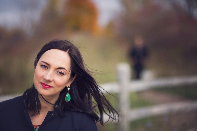 A woman stands for a portrait, her shoulders and head are visible in the bottom left of the frame. Her hair is blowing. In the background, out of focus, is a wooden fence. 