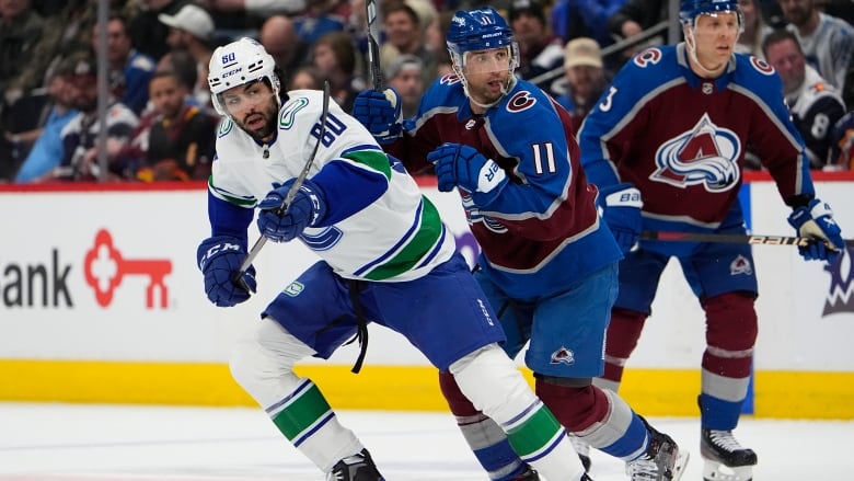 Three hockey players tussle on the ice, with a crowd behind them.