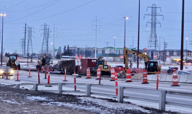 Machinery, including a backhoe, is seen on a bridge. The equipment and crews are surrounded by orange construction cones.