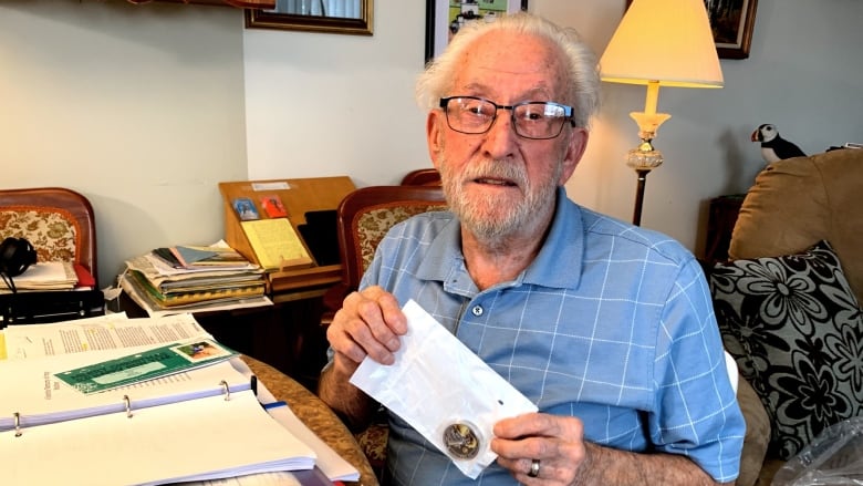 An elderly man with grey hair and wearing glasses is holding onto a clear plastic bag containing a silver coin. 