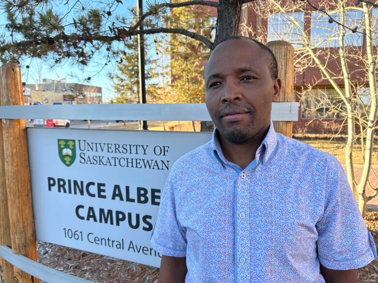 A Black man stands outside in front of a sign that says 'University of Saskatchewan Prince Albert Campus.'