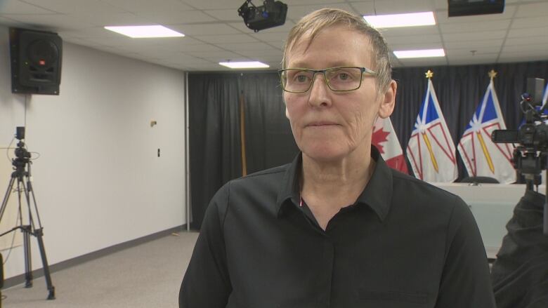 A woman with short hair and glasses wearing a black shirt stands in a conference room.