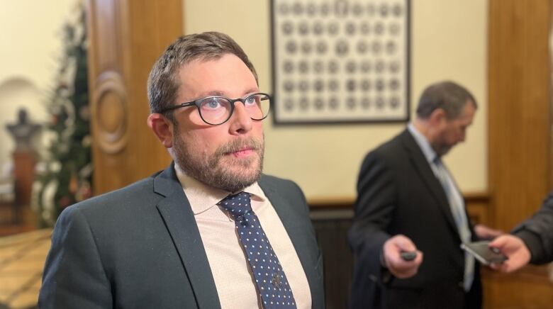 A man in a suit with glasses and a short beard in front of microphones.