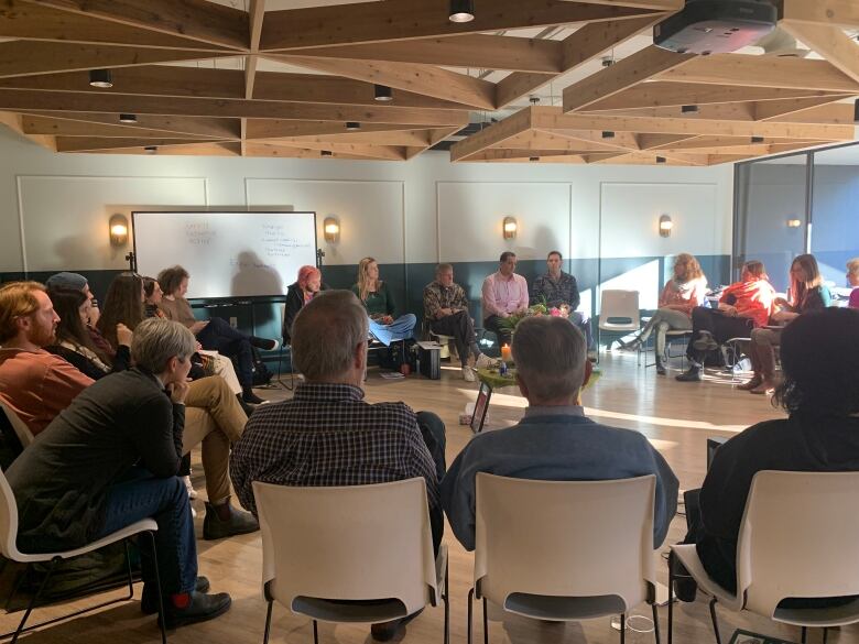 People sit in a circle on white chairs in a well-lit room. 