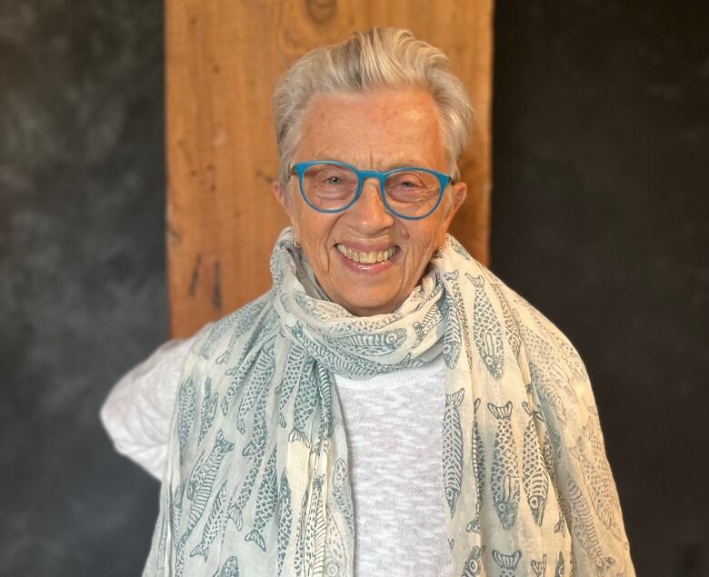 A Caucasian woman with blue glasses and short grey hair stands in front of a wooden column. 