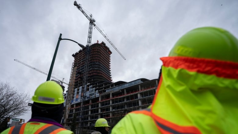 People in safety vests stare up at an under-construction building with a tower crane next to it.