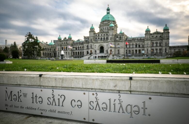 A signage with a subtitle that reads 'We all love the children (every child matters)', along with its Indigenous translation as the main title text in a larger font, is seen in the foreground. In the background, the B.C. Legislature in Victoria is pictured. 