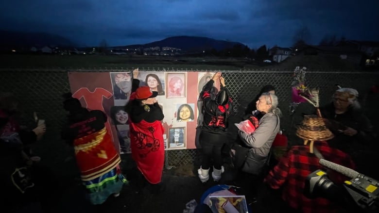 People put up posters and signs of women, along with bouquets and flowers, on a chain link fence on a dark evening.