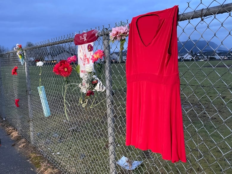 A red dress, bouquets and a sign honouring Missing and Murdered Indigenous women hang on a chain link fence.