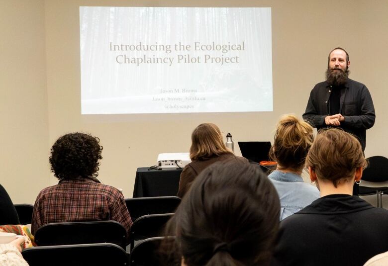 A man with a beard stands in front of people in chairs with a slide in the background that says 'Introducing the ecological chaplaincy pilot project.'