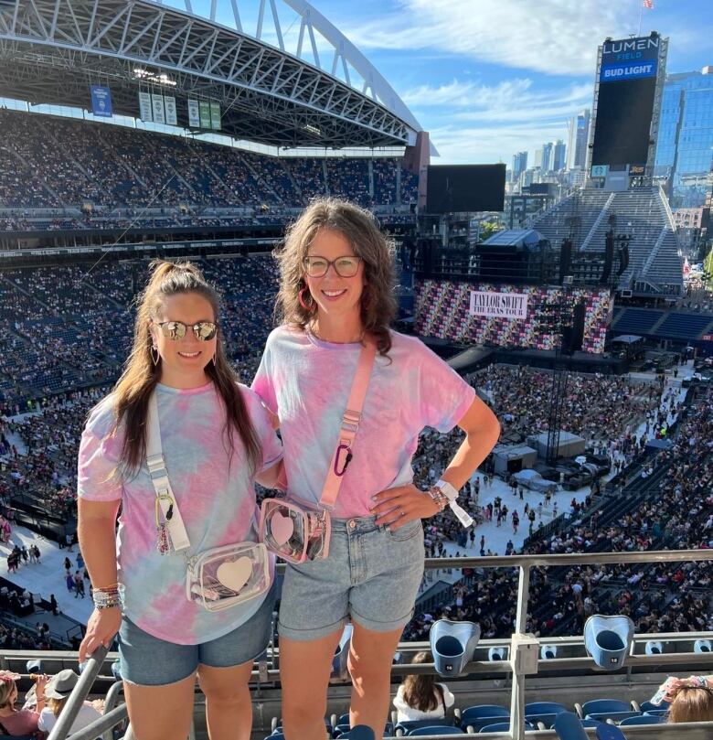 Two women in matching denim shorts and pink and blue tie dye t-shirts stand in a large concert arena with the stage in the background. 