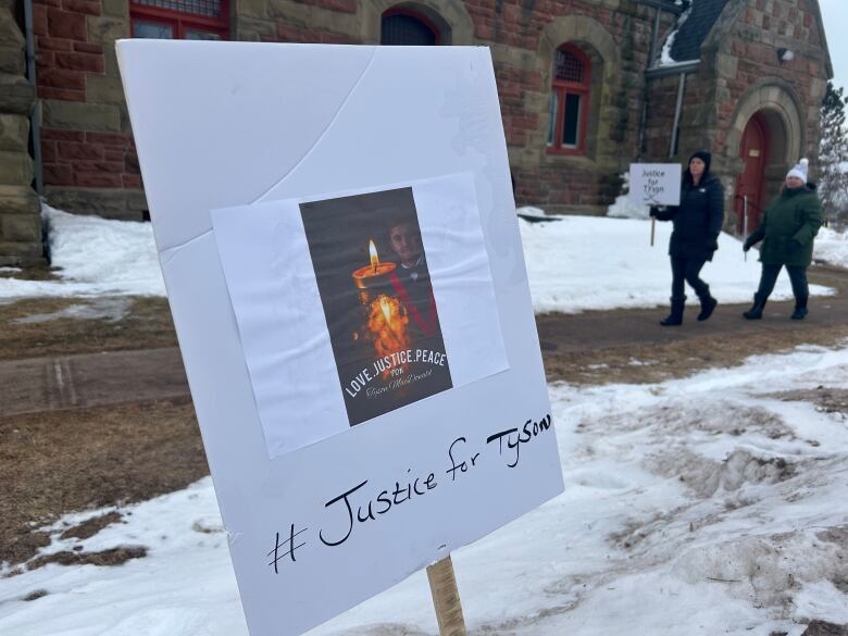 A sign reading Justice for Tyson stuck into the ground in front of a courthouse with people carrying placards in the background.