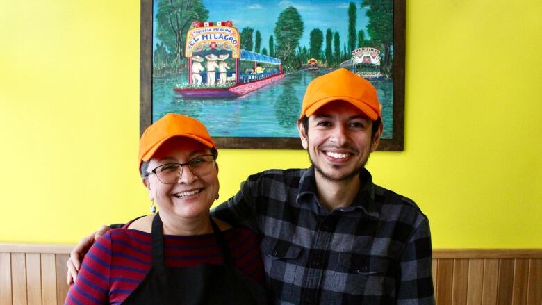 A young man stands with his arm around the shoulders of middle-aged woman. They are in a restaurant dining room paimnted yellow, in front of a paintaing of boats on a river.