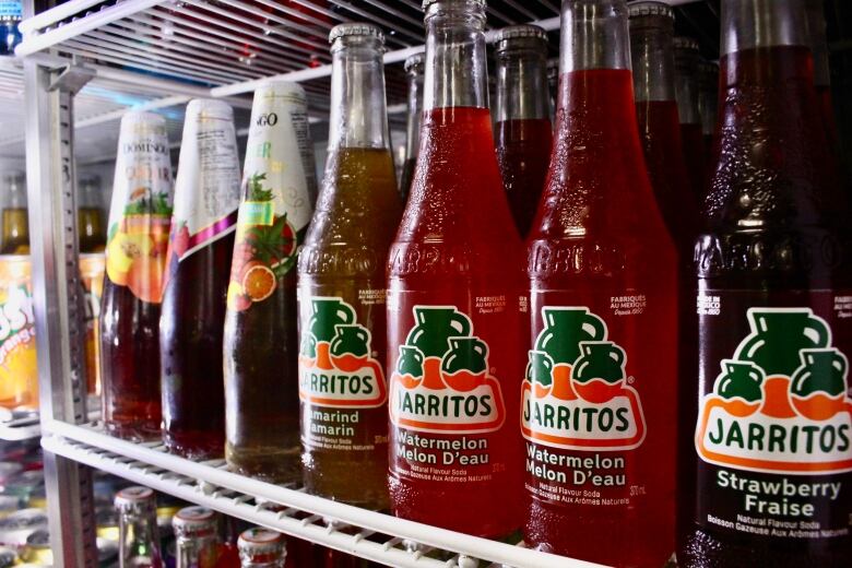 Different varieties of glass soda bottles sit on a shelf in a fridge. Four are the brand 'Jarritos' and are in the flavours tamarind, watermelon and strawberry.