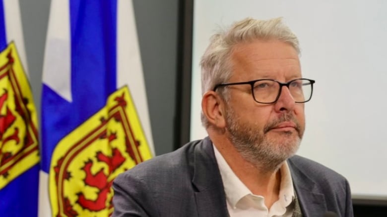 A man with white hair and wearing a grey suit sits in front of a Nova Scotia flag.