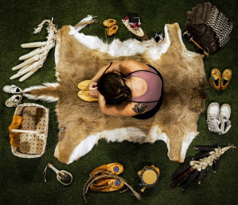 An Indigenous woman on a deer hide surrounded by cultural items like ash baskets and moccasins 