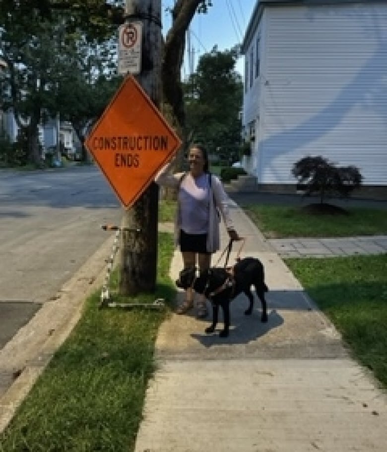 A white woman in a sweater and shorts stands beside a power pole with an orange diamond-shaped sign reading construction ends