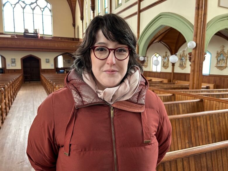 A woman with dark hair and glasses wearing a parka stands in a church. 