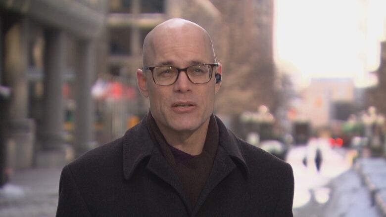 A man with shaven head and glasses, dressed in a dark woollen coat, stands on an Ottawa street corner.