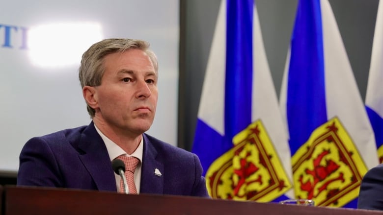 A man in a suit and tie sits near Nova Scotia flags.