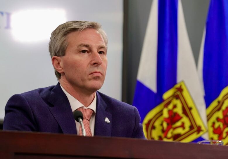 A man in a suit and tie sits near Nova Scotia flags.