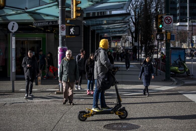 A person with a yellow hooded sweatshirt and an olive jacket is seen riding an e-scooter on a busy street.