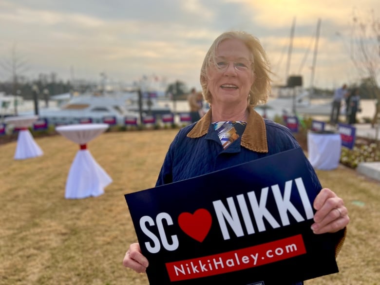 A blond woman with glasses holds a Nikki Haley sign while standing at a campaign event.