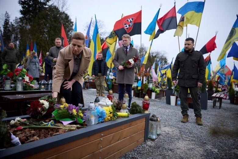 A woman in a winter coat lays down flowers at a monument as two men look on in a outdoor photograph. Behind them are a series of flags.