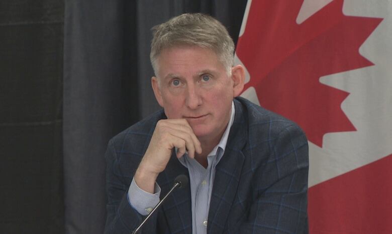 A middle-aged man, in a suit sits at a long table with a row of Newfoundland and Labrador and Canadian flags behind him.