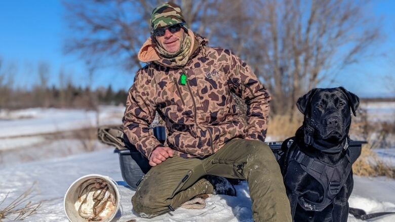 A man in outdoor wear poses with his black lab and a bucket of smelts 