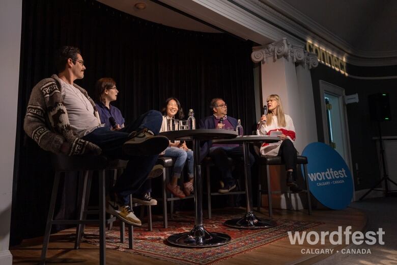 A tall brown haired man, a brown haired woman, an Asian woman, an Indian man, and woman with long blonde hair talk and laugh at two tables while holding mics. 