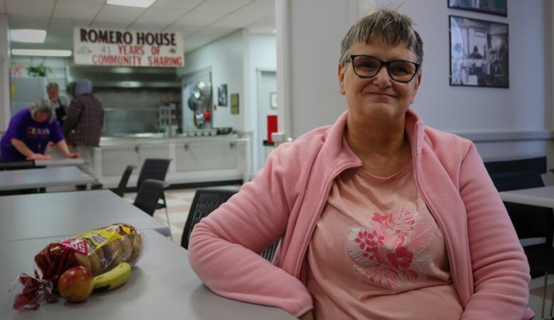 A smiling woman with short grey hair and a pink sweater sits in a cafeteria with a banana, bagels, and an apple. 