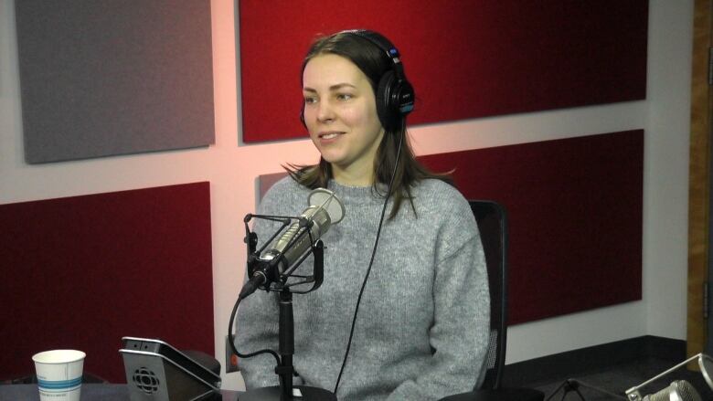 Woman with shoulder length brown hair in grey sweater sitting in front of a microphone.