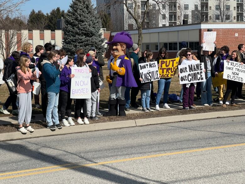 A group of demonstrators carrying signs, with a school mascot in the middle.