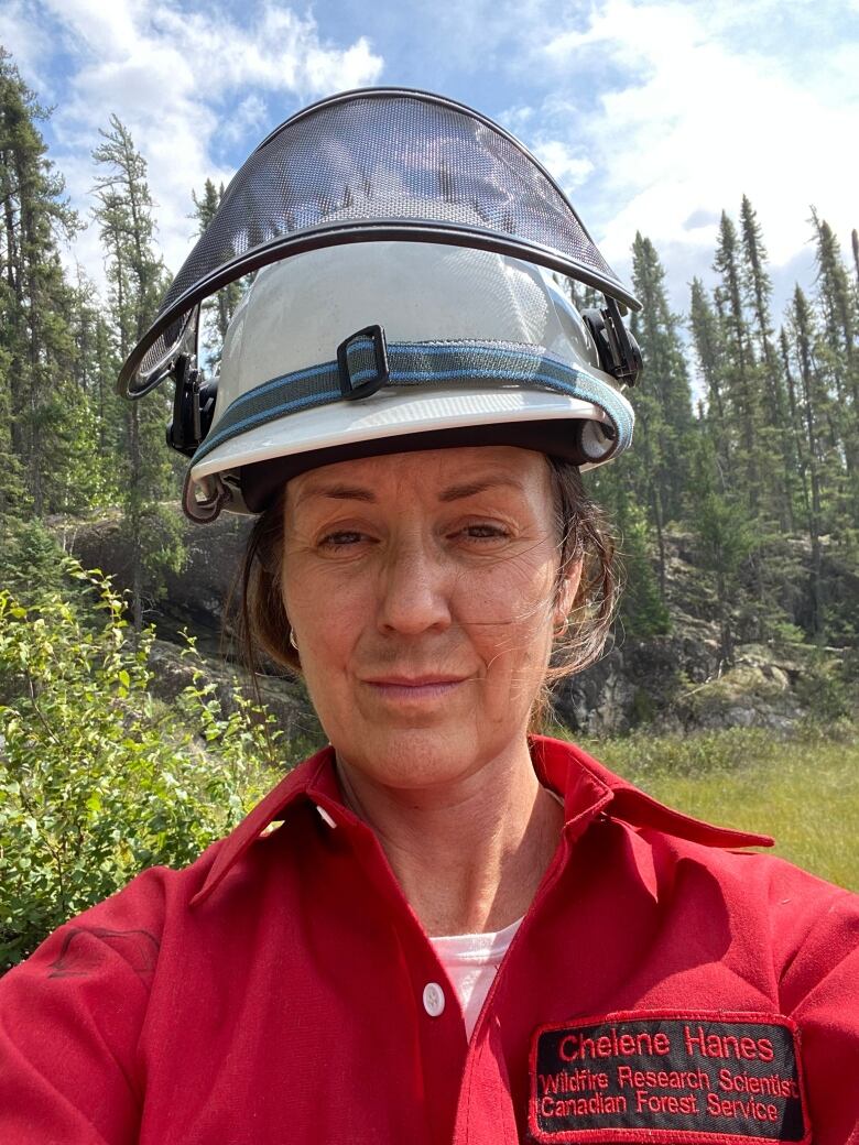 A woman in a forest wearing a firefighters helmet and red uniform.