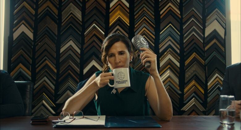 Michaela Watkins as Suze holds a coffee mug and a fan in front of a wall of frames.