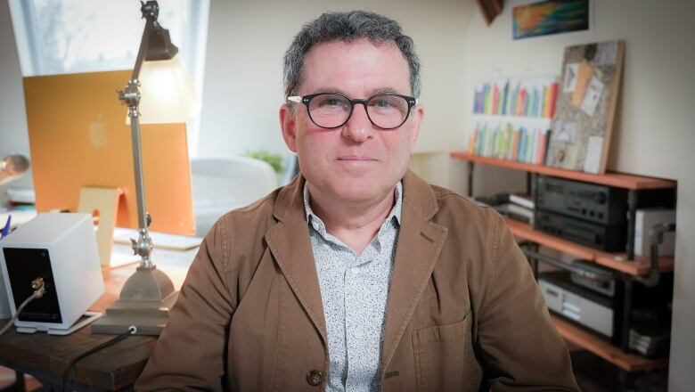 A man in a brown jacket and glasses smiles slightly at the camera while sitting in a bright indoor room, with a computer, artwork and stereo equipment behind him.