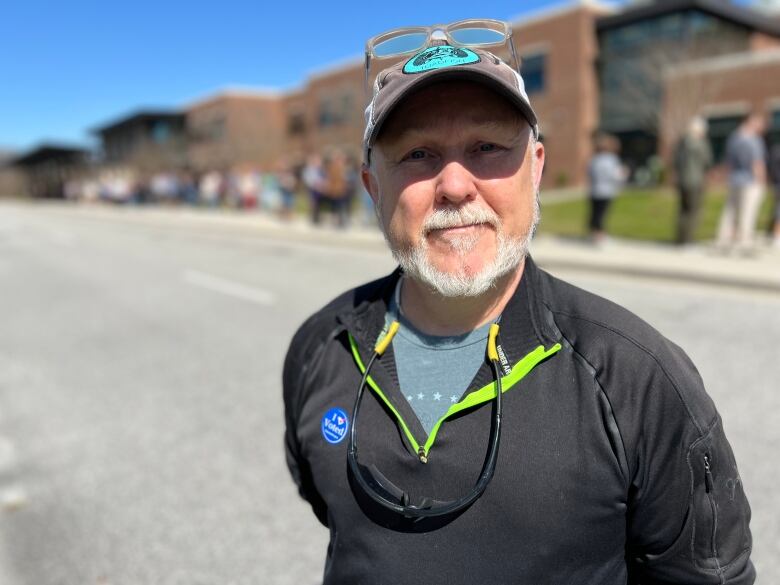 Man in baseball cap stands outside polling station, with long line in back