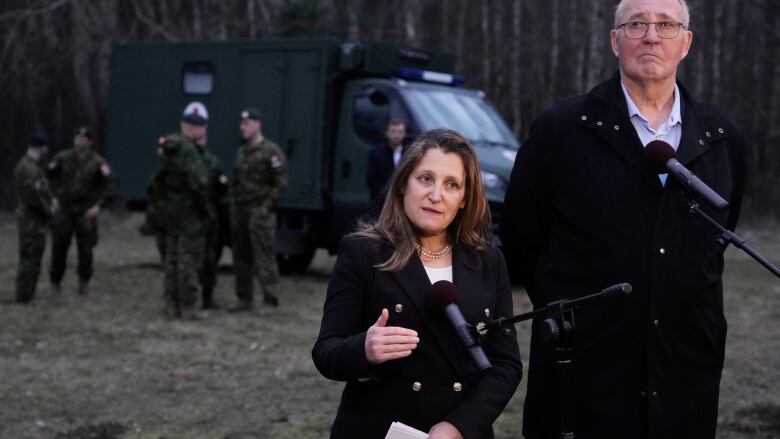 Two people speak at a microphone, with military medics in the background.