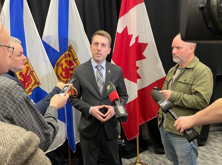 A man in a suit and tie stands in front of flags as people stand around him.