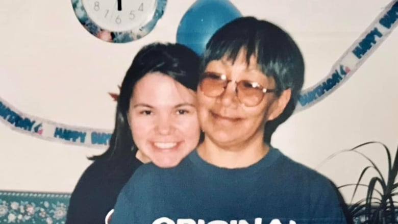 a woman rests her head on her mom's shoulder. They both smile at the camera. Balloons and a happy birthday banner hangs behind them. 