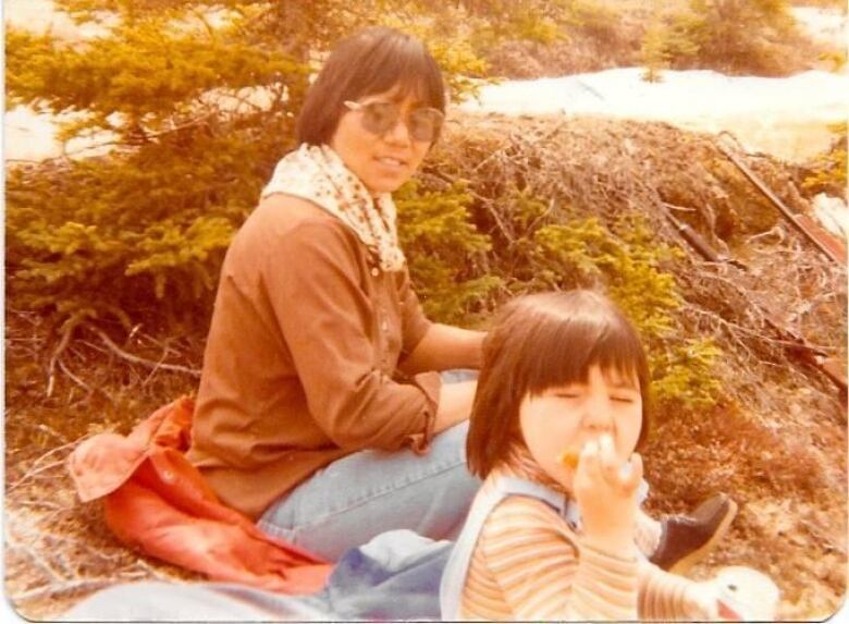 A photo from 1980 with a mother and a daughter eating food while sitting in the grass. 