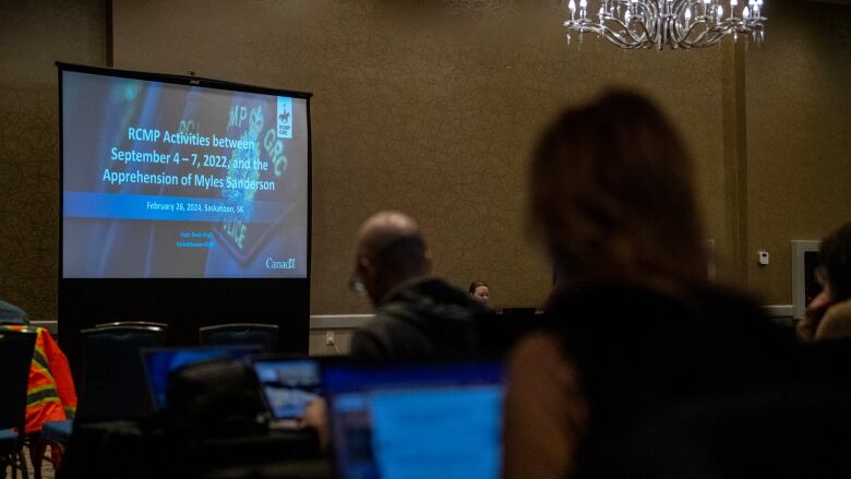 A screen shows the title page of a presentation on RCMP activity before the arrest of Myles Sanderson. Several people in the foreground are out of focus.