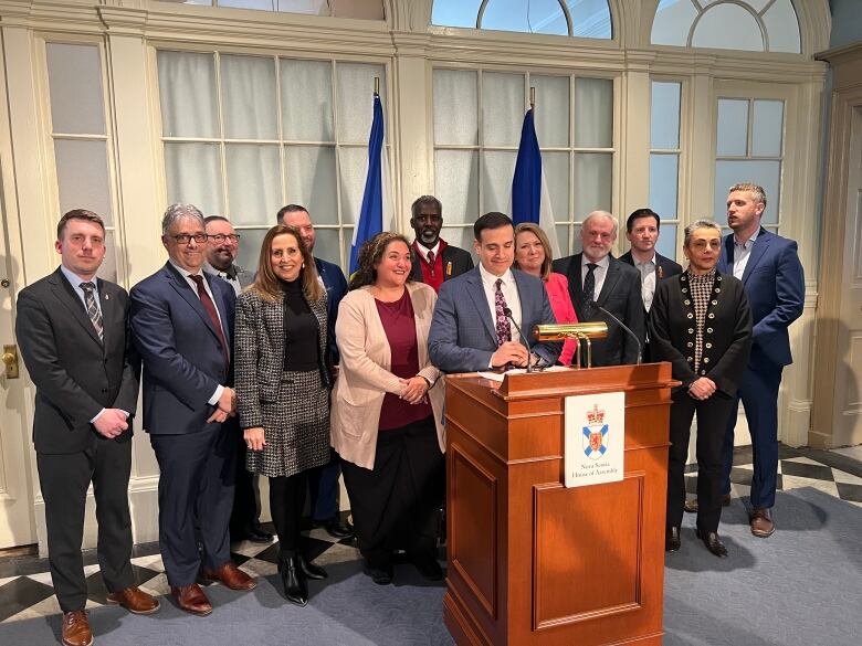 A group of people are seen standing around a man in a blue suit at a podium