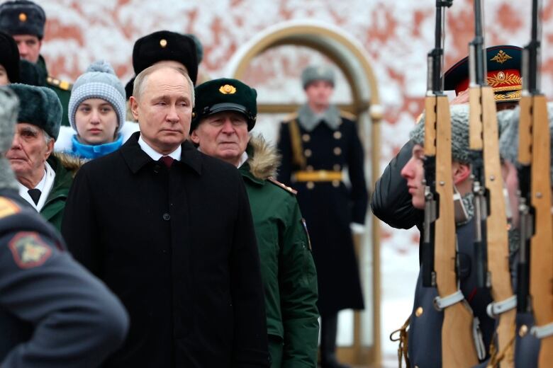 A man in a long black jacket, over a white shirt and dark red tie, stand among other people outside next to a line of soldiers holding rifles upward. 