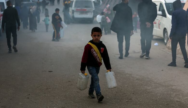 A young child wearing a red hoodie carires two white cans of fuel. Around him are people and cars.