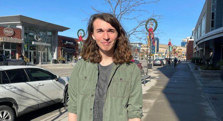 A university student with shoulder-length brown hair and a gray t-shirt worn underneath a green jacket stands on a street in Uptown Waterloo, Ontario.