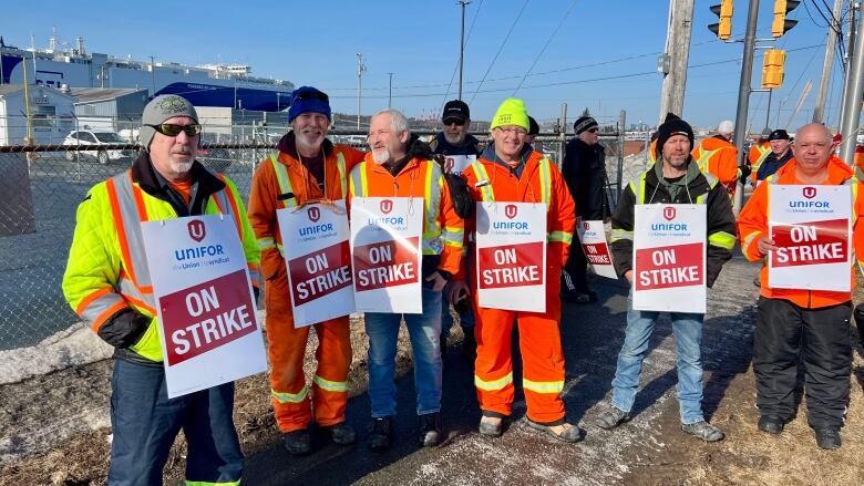 Six workers hold signs that say 'on strike' in front of a port with a large boat docked. 